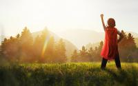 Self-Determination in Coaching To the right of the picture stands a young boy, with his back to the camera, standing with his legs apart, right hand on his hip. His left hand is in a fist and raised towards the sky. He is wearing a white shirt, jeans, and a red cape.